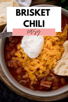 a bowl filled with chili and cheese on top of a wooden table next to bread