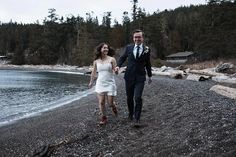 a man and woman holding hands walking on the beach next to the water with trees in the background