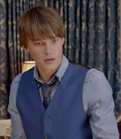 a young man wearing a blue vest and tie in front of a window with curtains