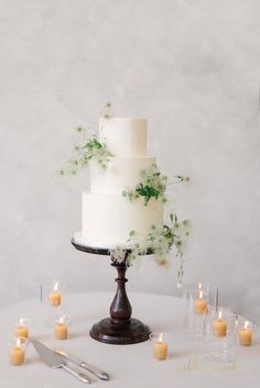 a white wedding cake with greenery on top surrounded by candles and silverware in front of a gray wall