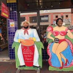 two people are standing next to giant statues in front of a storefront, one is holding a bucket and the other has a sign that says little havana market place