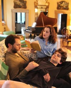 two men and a woman sitting on a couch in a living room with piano behind them