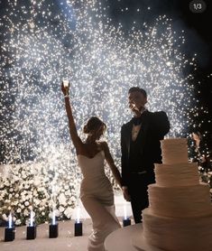 a newly married couple standing next to a wedding cake with fireworks in the background at night