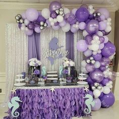 a purple and white dessert table with mermaid balloons on the wall, under an arch