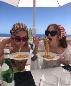 two women sitting at an outdoor table with plates of spaghetti in front of their mouths