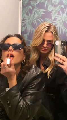 two women in sunglasses are brushing their teeth while taking a selfie with an iphone