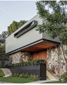 a modern house with stone walls and stairs leading up to the front door, surrounded by greenery