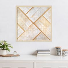 a white dresser with some books and plants on it in front of a wall mounted art piece