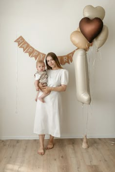 a woman holding a baby in front of balloons and a happy birthday banner on the wall