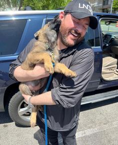 a man holding a dog in front of a blue truck with the door open and his tongue out