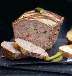 sliced meatloaf with pickles and bread on black slate board next to knife