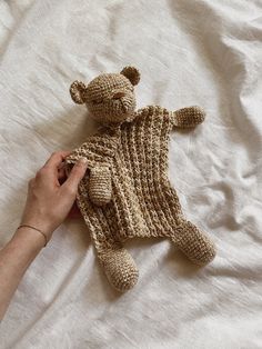 a person holding a knitted teddy bear on top of a white bedding sheet