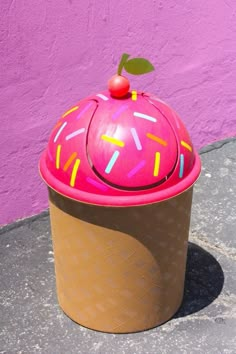 a cupcake shaped container with sprinkles and a cherry on top sits in front of a pink wall