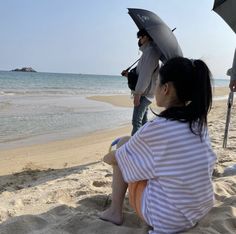 a woman sitting on top of a sandy beach next to the ocean holding an umbrella