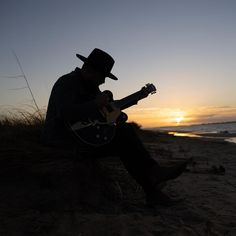 a man sitting on top of a sandy beach next to the ocean playing a guitar