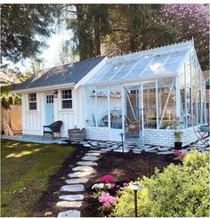 a small white house with a glass roof in the yard, surrounded by flowers and trees