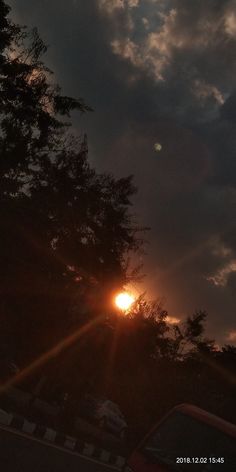 the sun is setting behind some trees in the dark sky with clouds and cars parked on the road