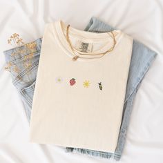 Welcome the sunny days with our Sunkissed Strawberry and Bee Embroidered T-Shirt! Made from 100% cotton Soft Comfort Colors, this shirt is both stylish and comfortable, perfect for your summer adventures.Features:Material: 100% cotton Soft Comfort ColorsSizes: S, M, L, XL, 2XL (refer to size chart in images)Colors Available: Banana, Bay, Blossom, Chalky Mint, Chambray, Ivory, Orchid, Peachy, Violet (Note: Colors may vary slightly due to monitor settings)Design: Summer-inspired embroidery featuring a daisy, strawberry, sun, and bee on the center chestCare Instructions: Machine wash cold, tumble dry lowThis Sunlissed Strawberry and Bee Embroidered T-Shirt is a delightful addition to your wardrobe, bringing a touch of sunshine wherever you go. Perfect for picnics, beach days, or casual outing Trendy Embroidered Summer T-shirt, Casual Embroidered Organic Cotton Tops, White Embroidered T-shirt For Summer, Cream Cotton Top With Floral Embroidery, Cute Spring Tops In Organic Cotton, Everyday White Embroidered T-shirt, White Embroidered T-shirt, White Embroidered T-shirt For Everyday, Cream Embroidered Cotton Tops