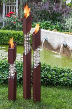 three tall metal sculptures with flames in the middle of grass and bushes behind them, on either side of a pond