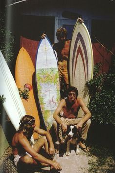 two men and a dog are sitting in front of surfboards with one man holding a dog