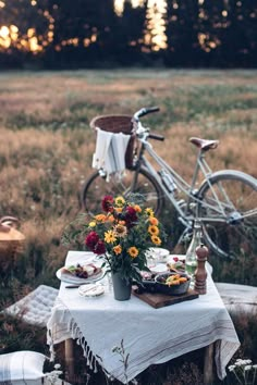 a table with food and flowers on it in the middle of a field next to a bicycle