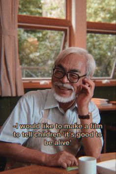 an older man sitting at a table talking on a cell phone with a cup in front of him