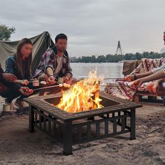 four people sitting around a fire pit in front of a body of water with tents