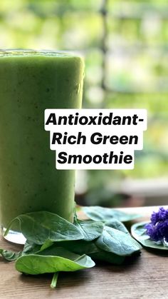 a green smoothie in a tall glass next to some leaves and purple flowers on a wooden table