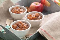 three small white bowls filled with apples on top of a cloth next to two spoons