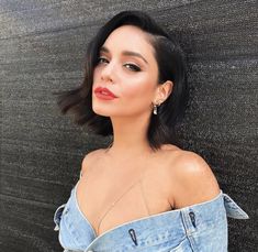 a woman with black hair wearing a denim dress and earrings, leaning against a wall