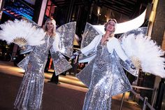 two women dressed in silver sequins and holding large white feathers are posing for the camera