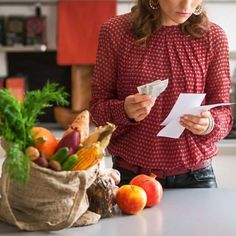 a woman standing at a kitchen counter holding a piece of paper and looking at her receipt