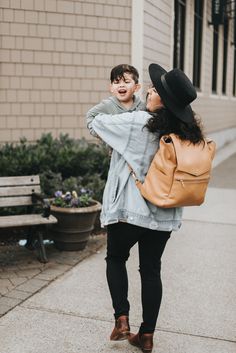 a woman carrying a young boy on her back while walking down the sidewalk in front of a building