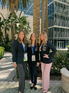 three women standing next to each other in front of a palm tree and glass building