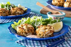 two plates with crab cakes and salad on blue table cloth next to utensils