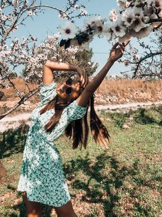 a woman in a green dress reaching up to the branches of a tree with white flowers