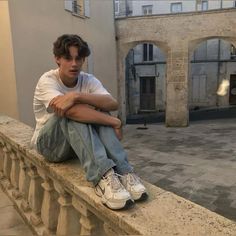 a young man sitting on top of a stone wall next to a building and looking off into the distance