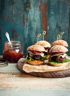 three hamburgers with meat and lettuce are on a wooden platter next to a jar of ketchup