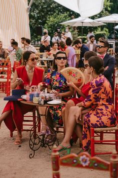 two women sitting at a table in front of other people, one holding a fan