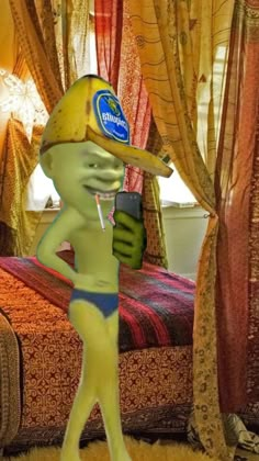 a green man in a yellow hat is holding a cell phone and standing next to a bed