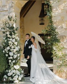 a bride and groom standing under an umbrella in front of a stone building with white flowers