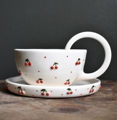 two white cups and saucers with cherries on them sitting on a wooden table