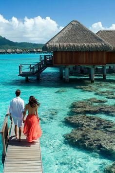 a man and woman are walking on a dock in the water near some huts with thatched roofs