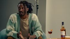 a man with dreadlocks sitting at a table next to a bottle of booze