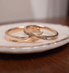 two gold wedding rings sitting on top of a white plate next to each other in front of a wooden table