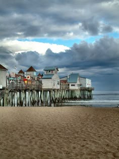 a pier on the beach with buildings in the background and people walking along it under a cloudy sky