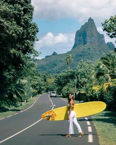 a man holding a yellow surfboard on the side of a road with mountains in the background