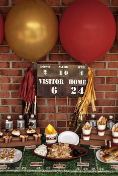 a table topped with lots of food next to red and gold balloons in front of a brick wall