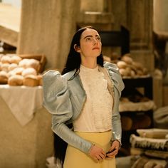 a woman with long black hair wearing a white blouse and yellow skirt standing in front of bread