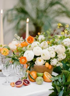 an arrangement of flowers, fruit and candles on a white table cloth with greenery